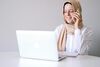 Woman wearing a beige hijab and white shirt smiling while talking on the phone and working on a laptop at a white desk