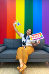 Lady sitting on sofa infront of rainbow wall holding happy pride signs 