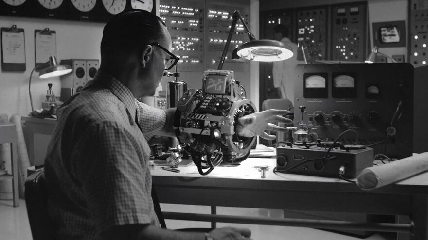 Man at a desk with various inventions around him in black and white