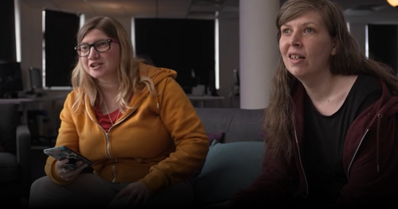 One woman holding a phone in her hand, sitting on a couch next to a second woman holding a video game controller.
