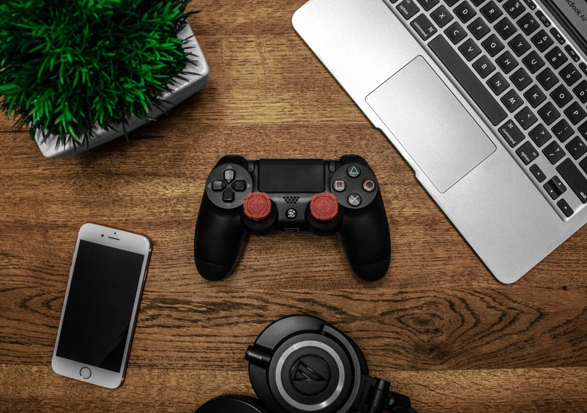 Gaming controller, smartphone, headphones, and laptop on a wooden desk.