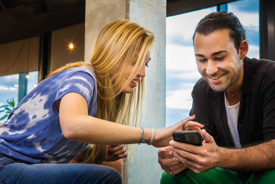 A man is looking at his phone while a woman reaches over to also see it, both are smiling. 